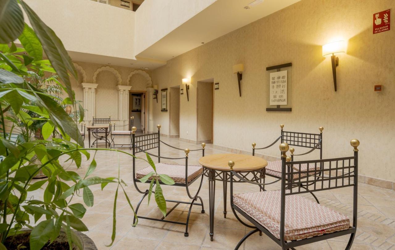 Hotel Ilunion Merida Palace Exterior photo The photo shows a cozy indoor seating area. There are a few metal-framed chairs with soft cushions arranged around a small round table. The walls are painted in a neutral color, and there are wall sconces providing warm lighting. In the background, y