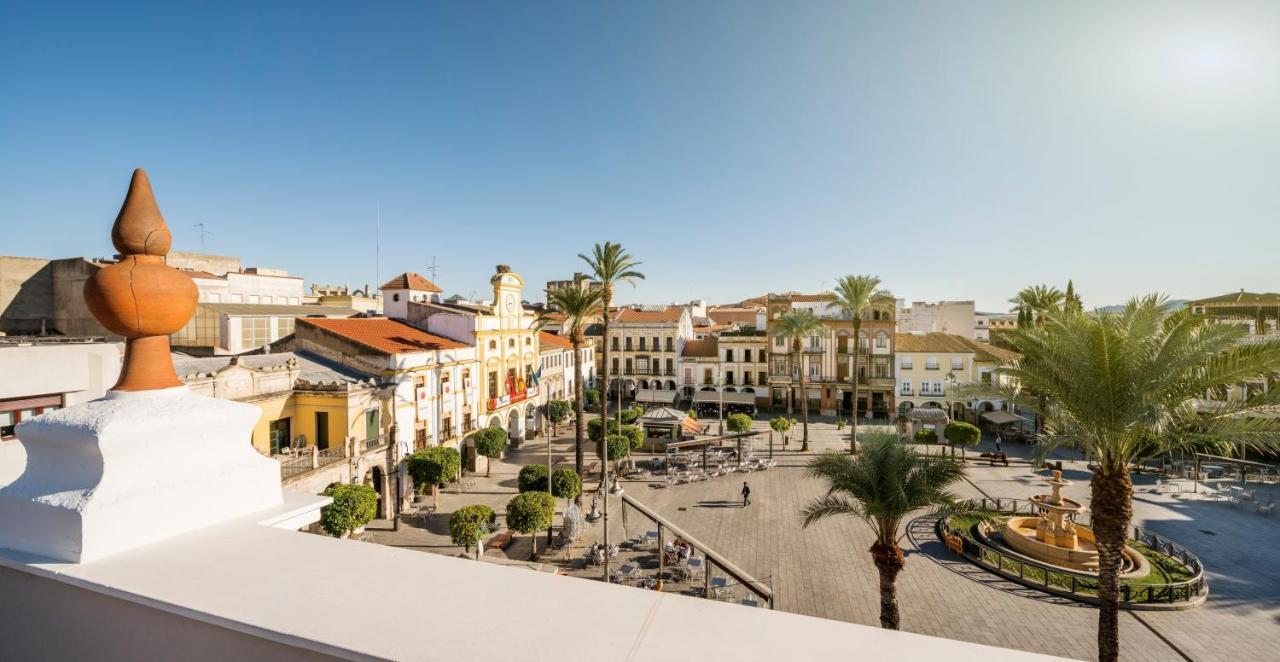 Hotel Ilunion Merida Palace Exterior photo The photo shows a picturesque town square or plaza with a sunny atmosphere. There are buildings with colorful facades lining the square, featuring traditional architectural elements. Palm trees are present, adding to the tropical feel. The square is 