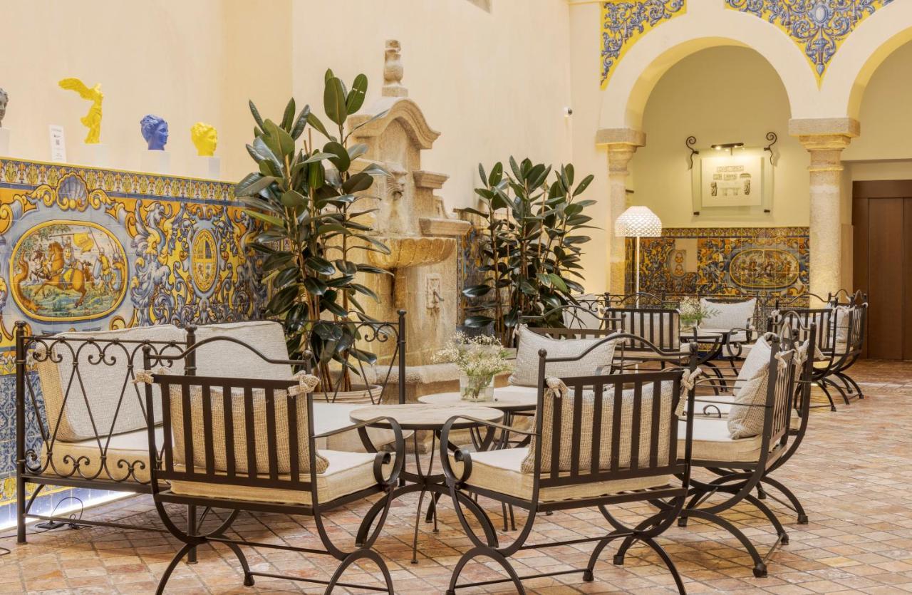 Hotel Ilunion Merida Palace Exterior photo The photo depicts a charming indoor space featuring a courtyard-like setting. It has several elegant wrought iron chairs and tables arranged neatly, suggesting a cozy gathering area. Lush green plants are positioned around the space, adding a touch o
