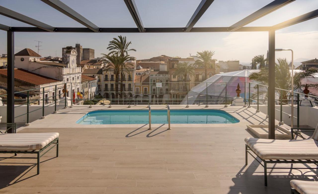 Hotel Ilunion Merida Palace Exterior photo The photo showcases a modern terrace with a swimming pool situated in a vibrant city setting. The terrace is framed by railings and has several lounge chairs placed nearby, suggesting a relaxing atmosphere. In the background, you can see a skyline th