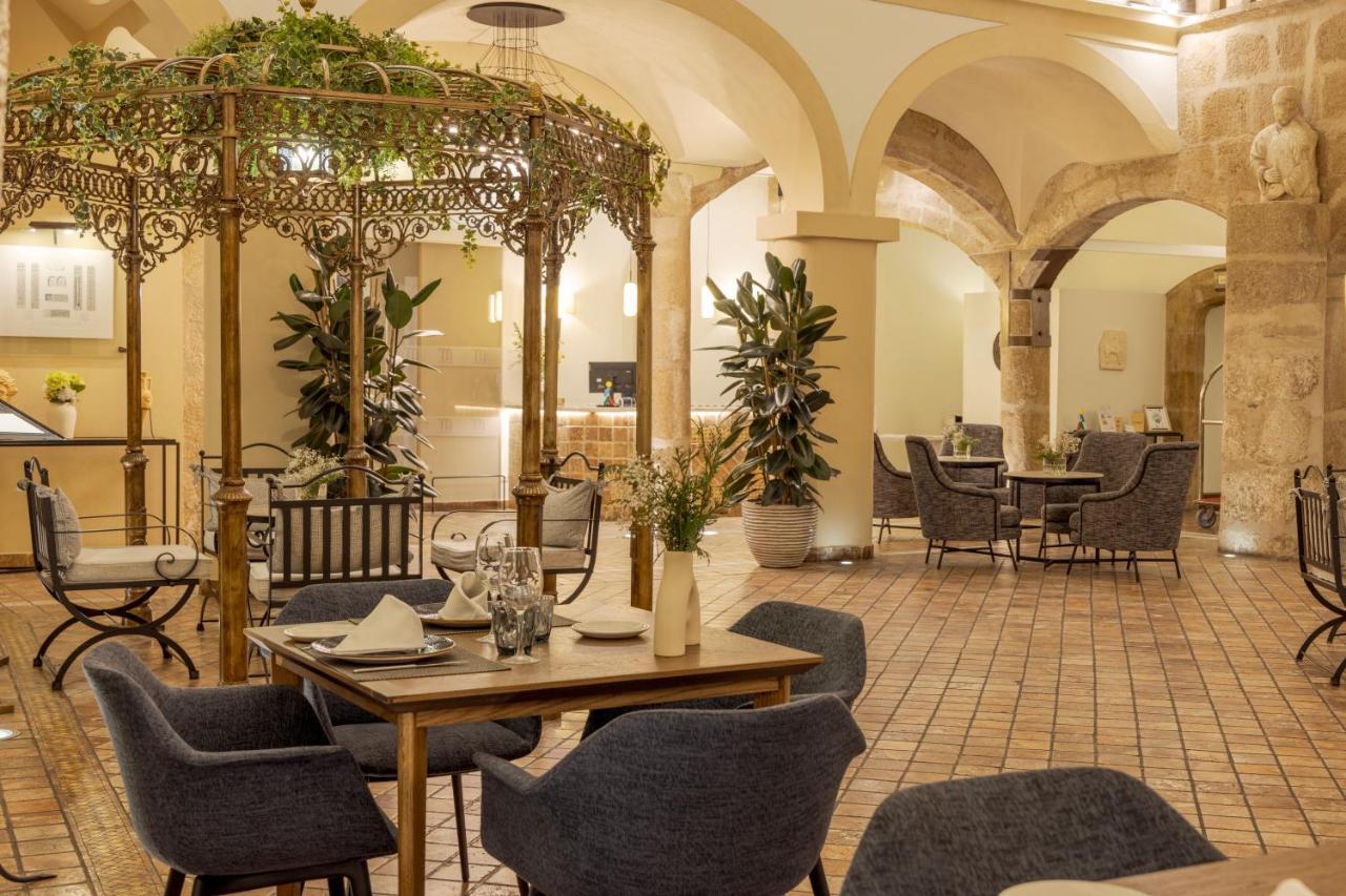 Hotel Ilunion Merida Palace Exterior photo The photo depicts an elegant indoor dining space with a charming atmosphere. In the center, there is a decorative gazebo-style structure adorned with greenery. Surrounding the gazebo are stylish tables with dark blue chairs, set for dining. The floor